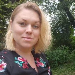 Portret kobiety z blond włosami w kwiecistej bluzce na tle zieleni.