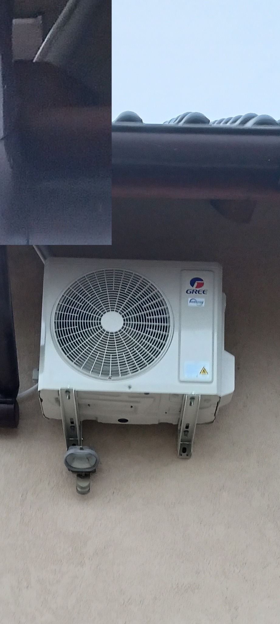 Zewnętrzna jednostka klimatyzacji marki Gree zamontowana na jasnej ścianie budynku z widocznym oświetleniem zewnętrznym pod spodem.