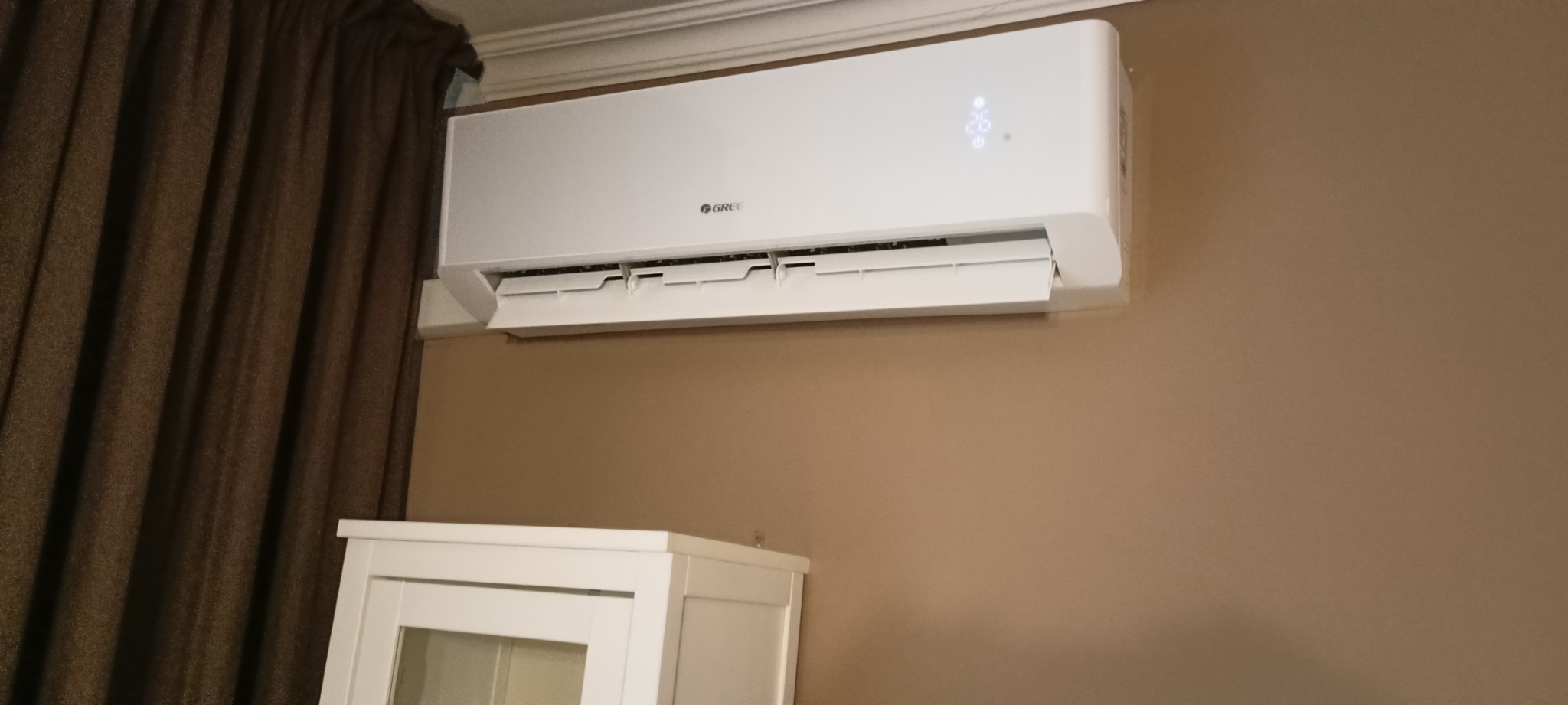 Biały klimatyzator marki Gree zamontowany na beżowej ścianie w pomieszczeniu z brązową zasłoną i białym meblem w tle.