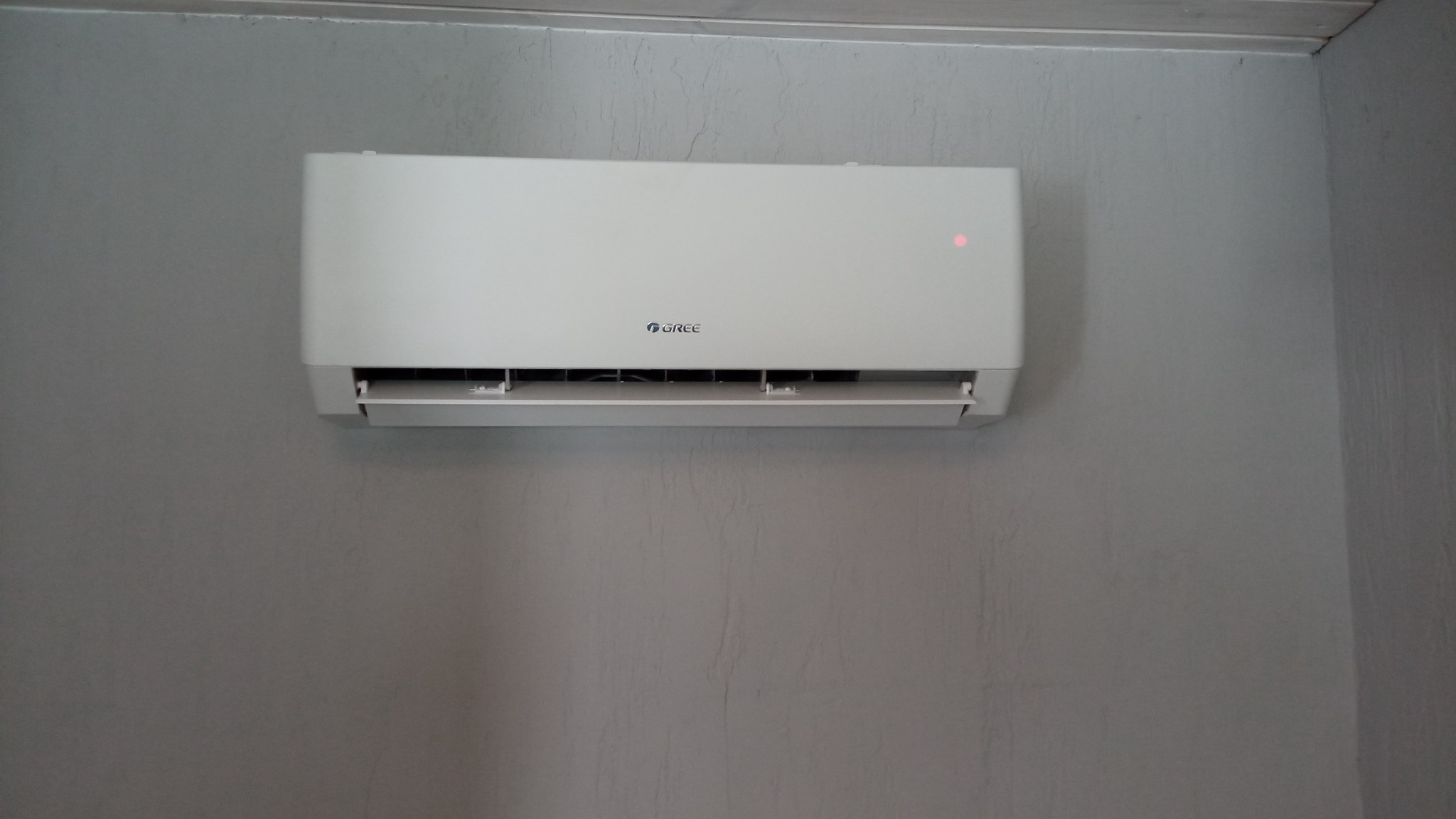 Biały klimatyzator marki Gree zamontowany na szarej, teksturowanej ścianie. Widoczna czerwona dioda sygnalizacyjna.
