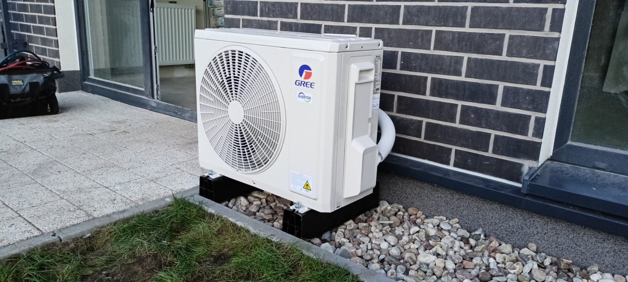 Zewnętrzna jednostka klimatyzacji Gree zamontowana na czarnych podstawkach, otoczona dekoracyjnymi kamieniami i trawą, przylegająca do ściany z ciemnej cegły i okna.
