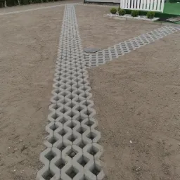 Ścieżka z ażurowych płyt betonowych prowadząca do domu z białym ogrodzeniem, teren wyrównany, częściowo przygotowany pod trawnik.