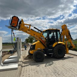 SZT Usługi - Żółta koparko-ładowarka JCB z osprzętem Catch Shift montuje prefabrykowane elementy betonowe na placu budowy. Widoczni pracownicy i przygotowane podłoże.