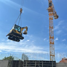 SZT Usługi - Żółta mini koparka podnoszona przez dźwig na tle nieba i szalunków fundamentowych. Widoczne zbrojenia i elementy konstrukcyjne.