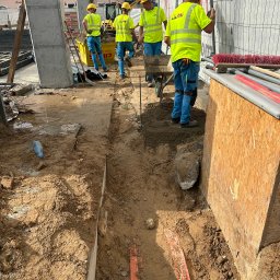 SZT Usługi - Pracownicy w żółtych kamizelkach układają pomarańczowe rury w wykopie. W tle widoczny sprzęt budowlany i fragmenty miejskiej zabudowy. Prace ziemne w toku.