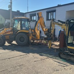 SZT Usługi - Żółta koparko-ładowarka JCB i minikoparka Yanmar w trakcie prac ziemnych na ulicy. Widoczne gruz i cegły, sugerujące rozbiórkę. W tle budynki mieszkalne i błękitne niebo.