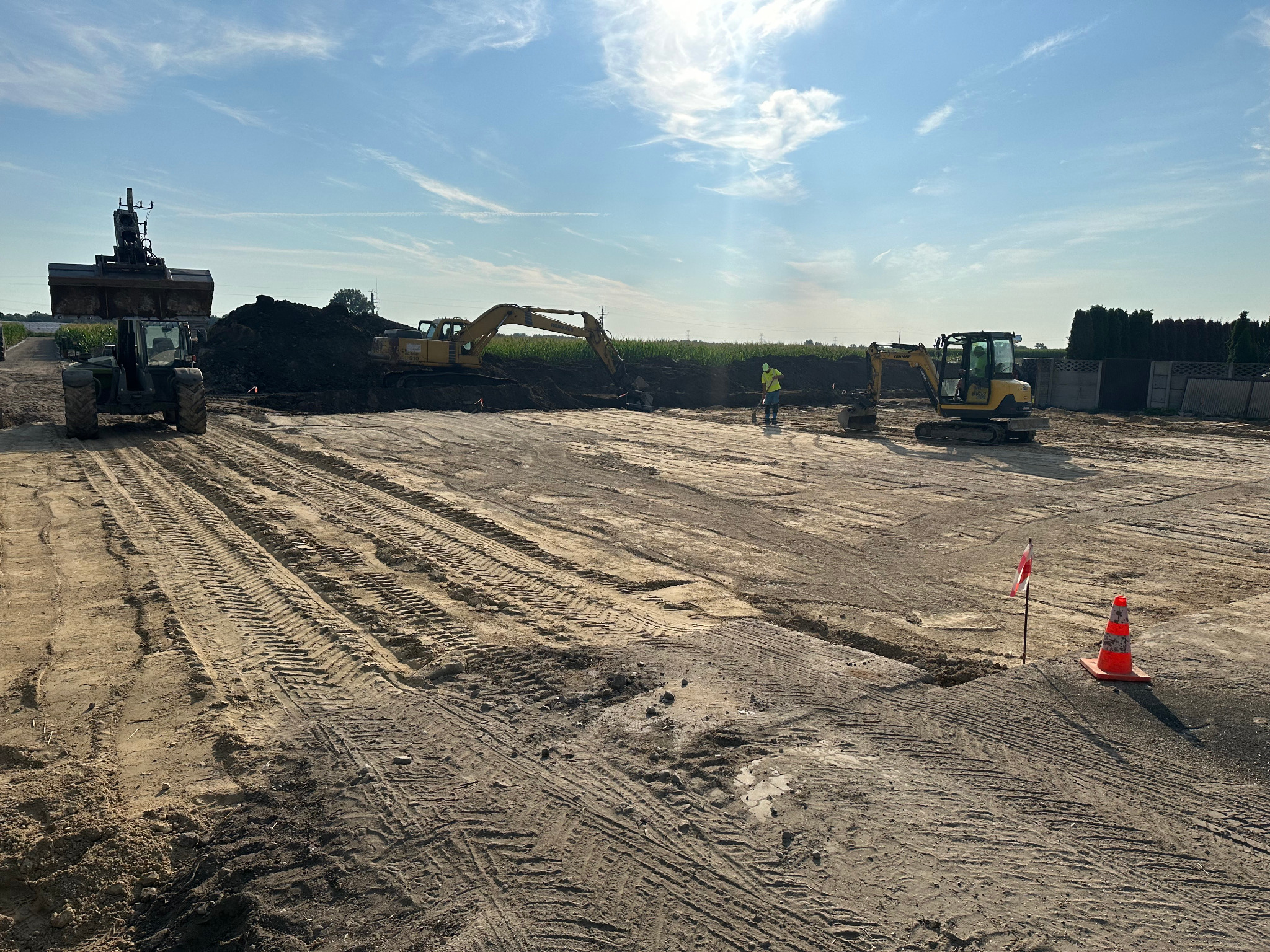 Niwelacja terenu z użyciem ciężkiego sprzętu: koparki, ładowarki. Widoczne ślady opon na wyrównanym terenie, pracownik w odblaskowej kamizelce i pachołek ostrzegawczy.