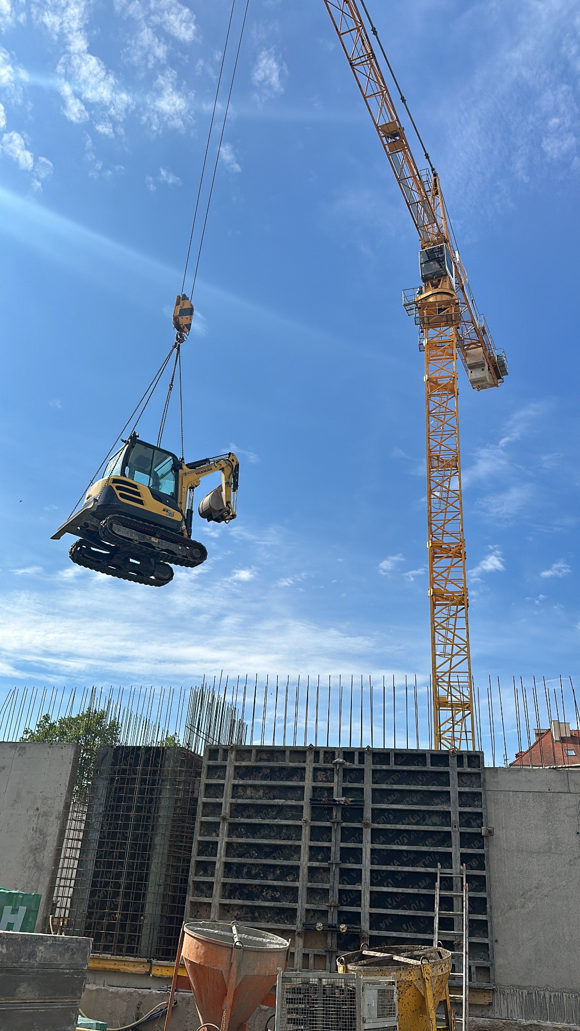 Żółta mini koparka podnoszona przez dźwig na tle nieba i szalunków fundamentowych. Widoczne zbrojenia i elementy konstrukcyjne.