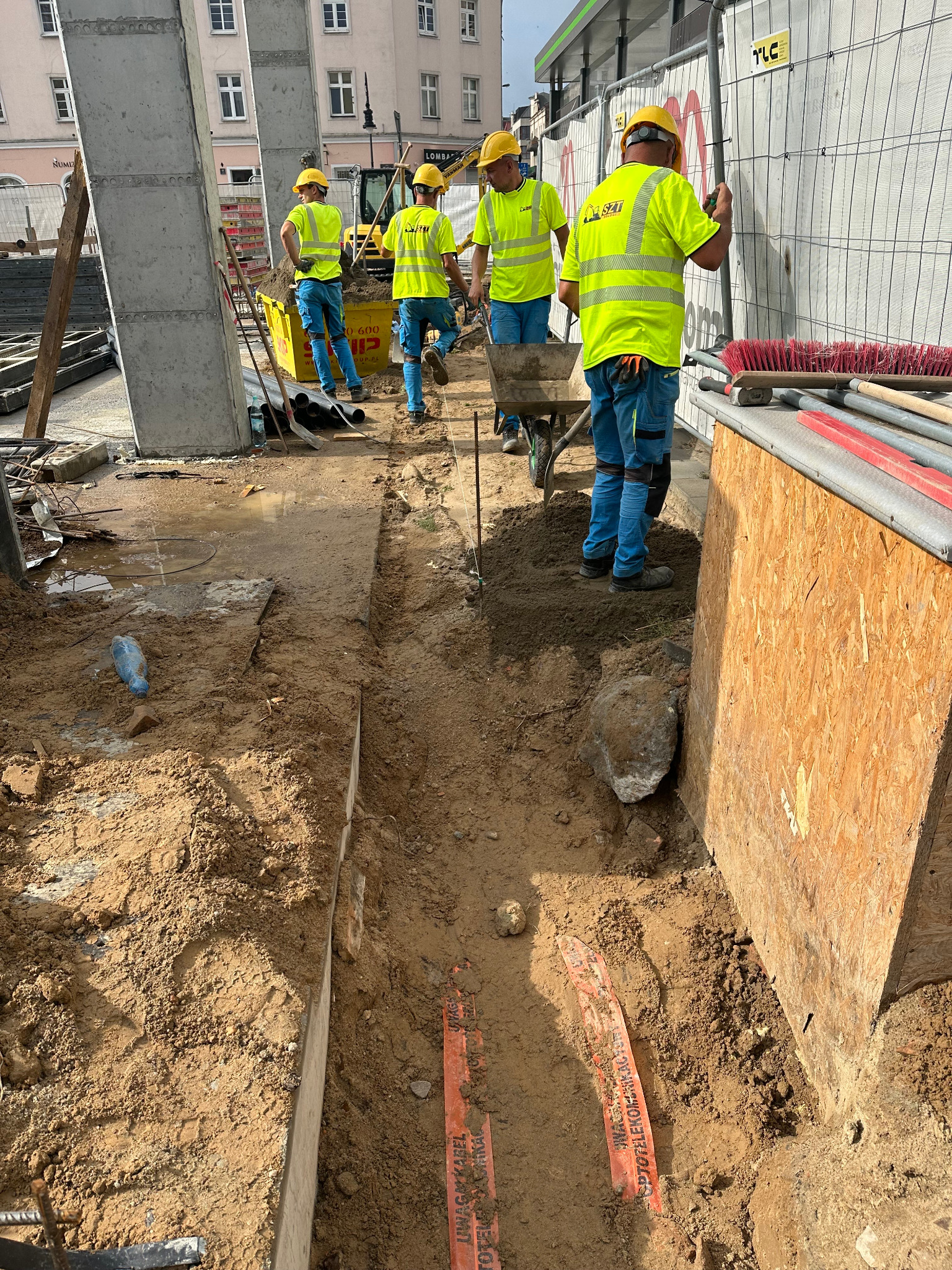 Pracownicy w żółtych kamizelkach układają pomarańczowe rury w wykopie. W tle widoczny sprzęt budowlany i fragmenty miejskiej zabudowy. Prace ziemne w toku.