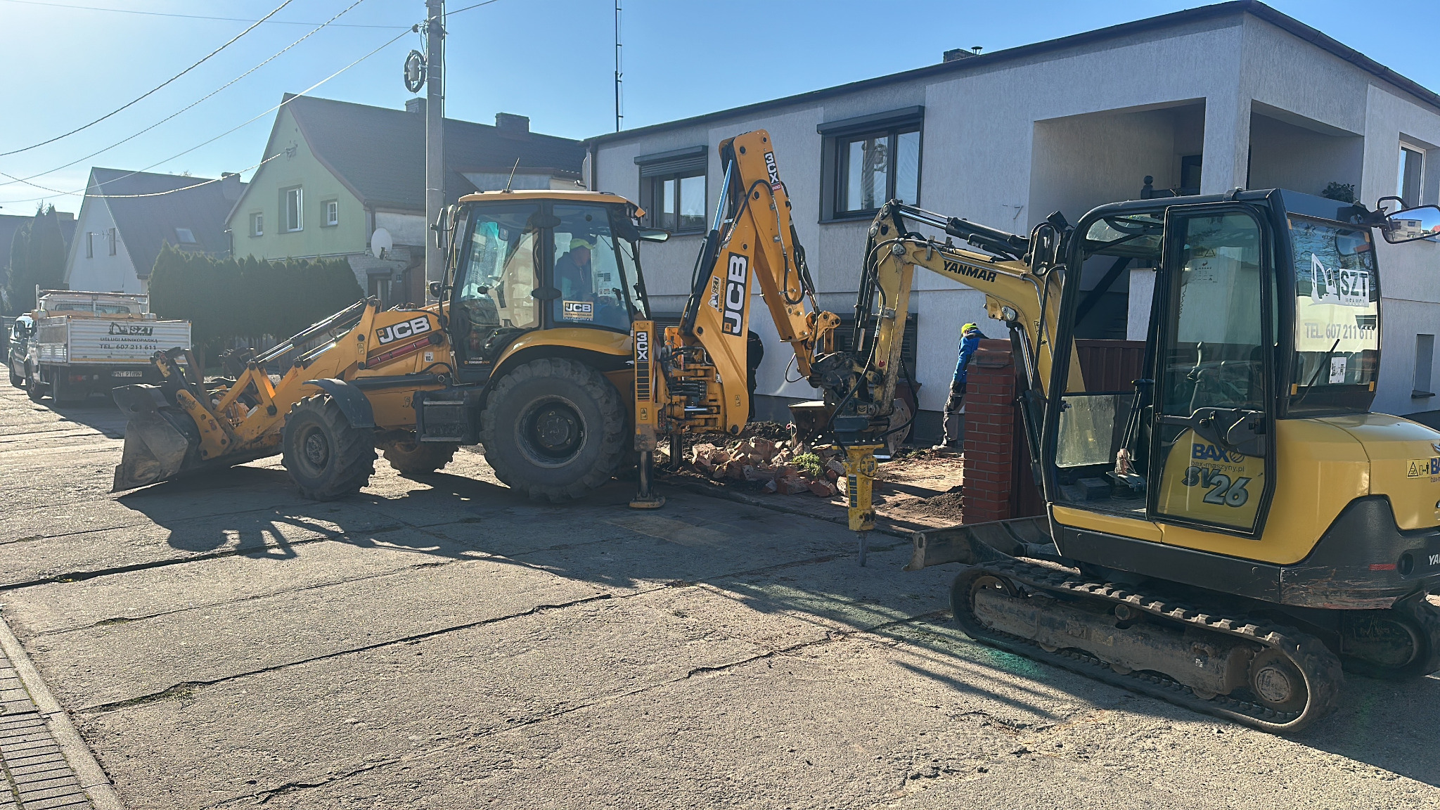 Żółta koparko-ładowarka JCB i minikoparka Yanmar w trakcie prac ziemnych na ulicy. Widoczne gruz i cegły, sugerujące rozbiórkę. W tle budynki mieszkalne i błękitne niebo.