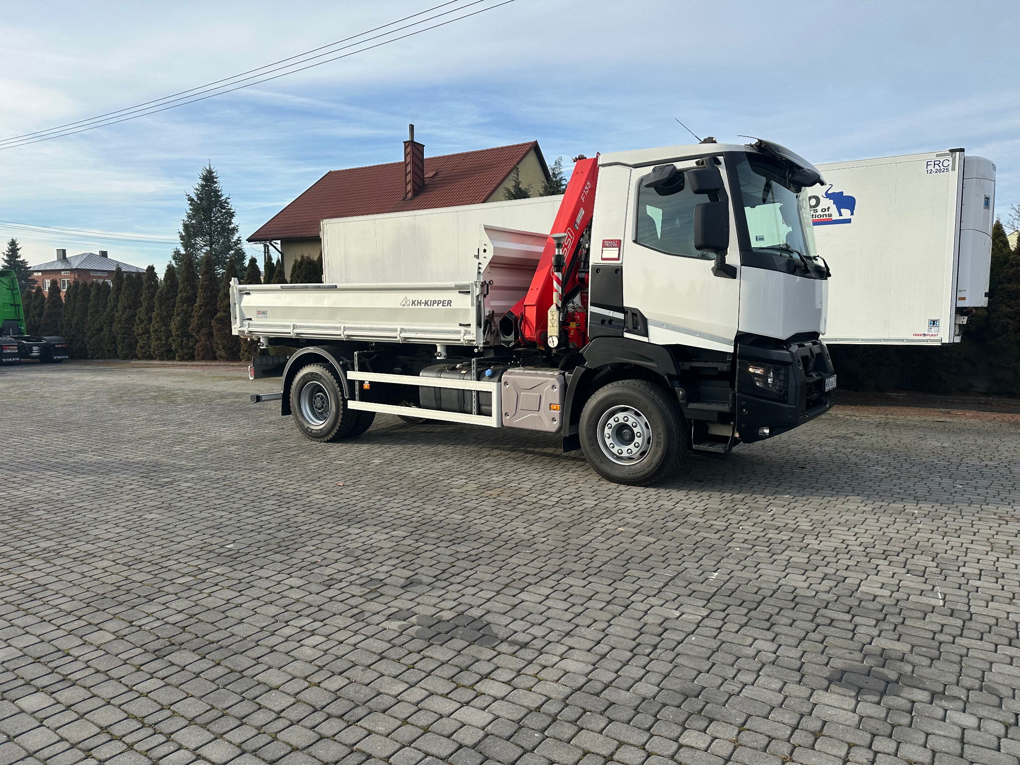 Ciężarówka z dźwigiem hydraulicznym KH-Kipper na kostce brukowej, z domem w tle. Widoczny czerwony dźwig i biała zabudowa skrzyniowa. Pojazd gotowy do transportu materiałów budowlanych.