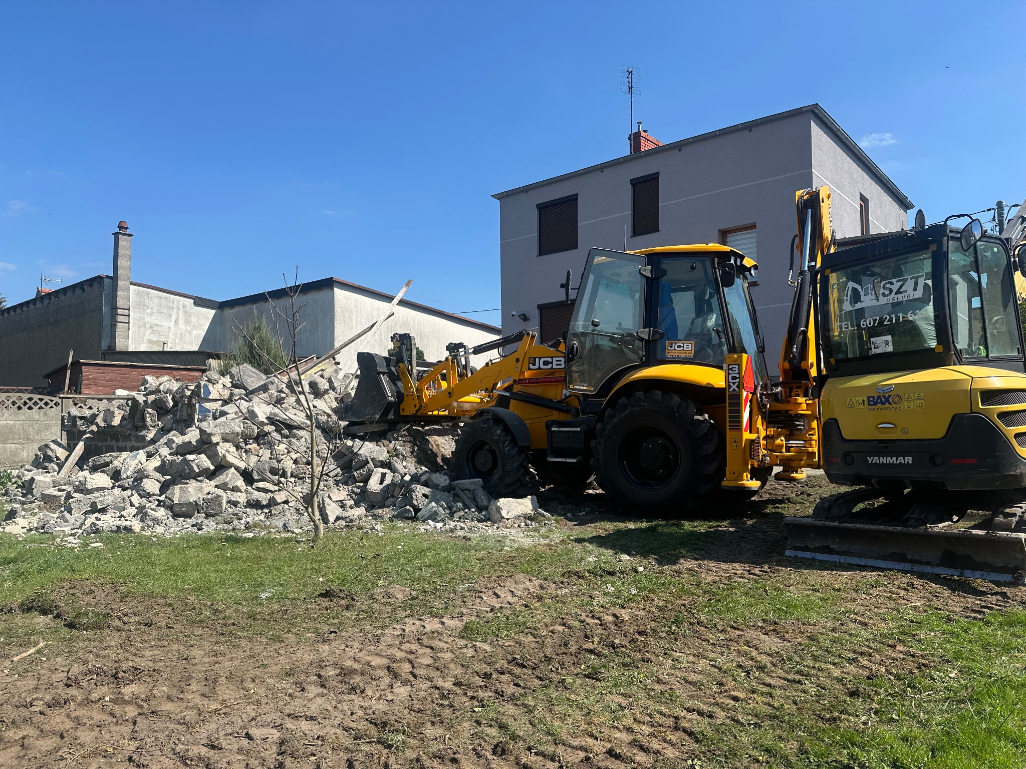 Żółta koparko-ładowarka JCB i minikoparka Yanmar na placu budowy z gruzem, w tle budynek mieszkalny. Widoczne ślady opon na trawie i ziemi.