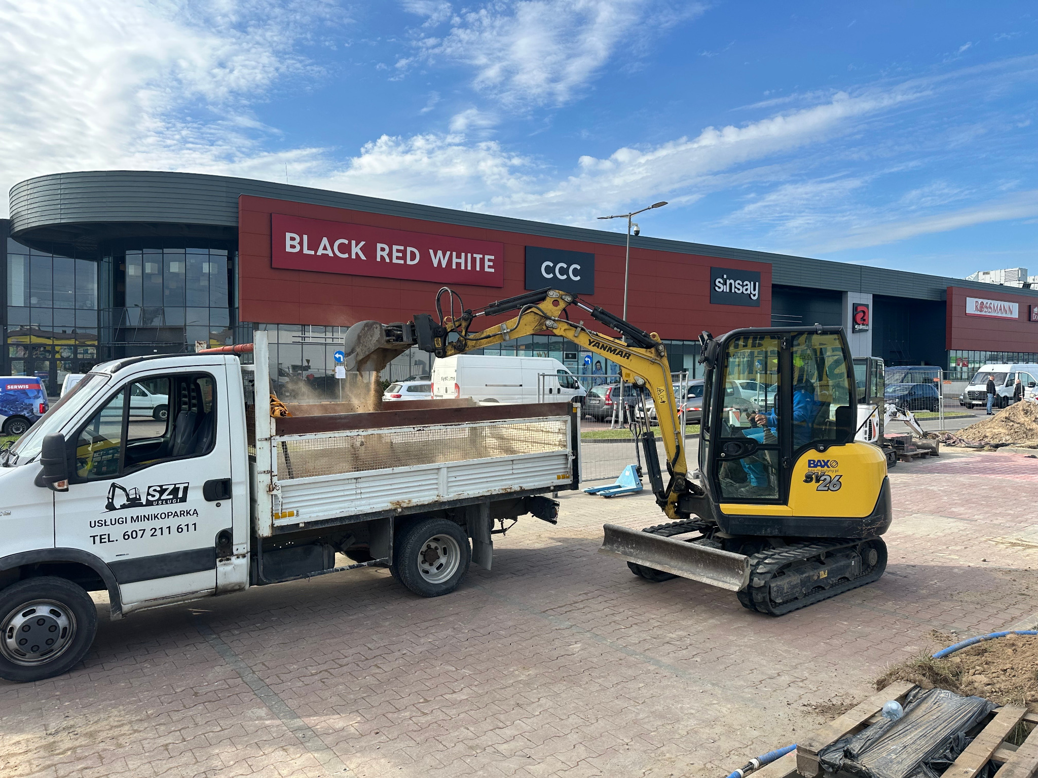 Żółta minikoparka Yanmar nabiera ziemię do skrzyni białej ciężarówki z logo firmy 'SZT Usługi Minikoparką' na tle budynku galerii handlowej z szyldami sklepów.