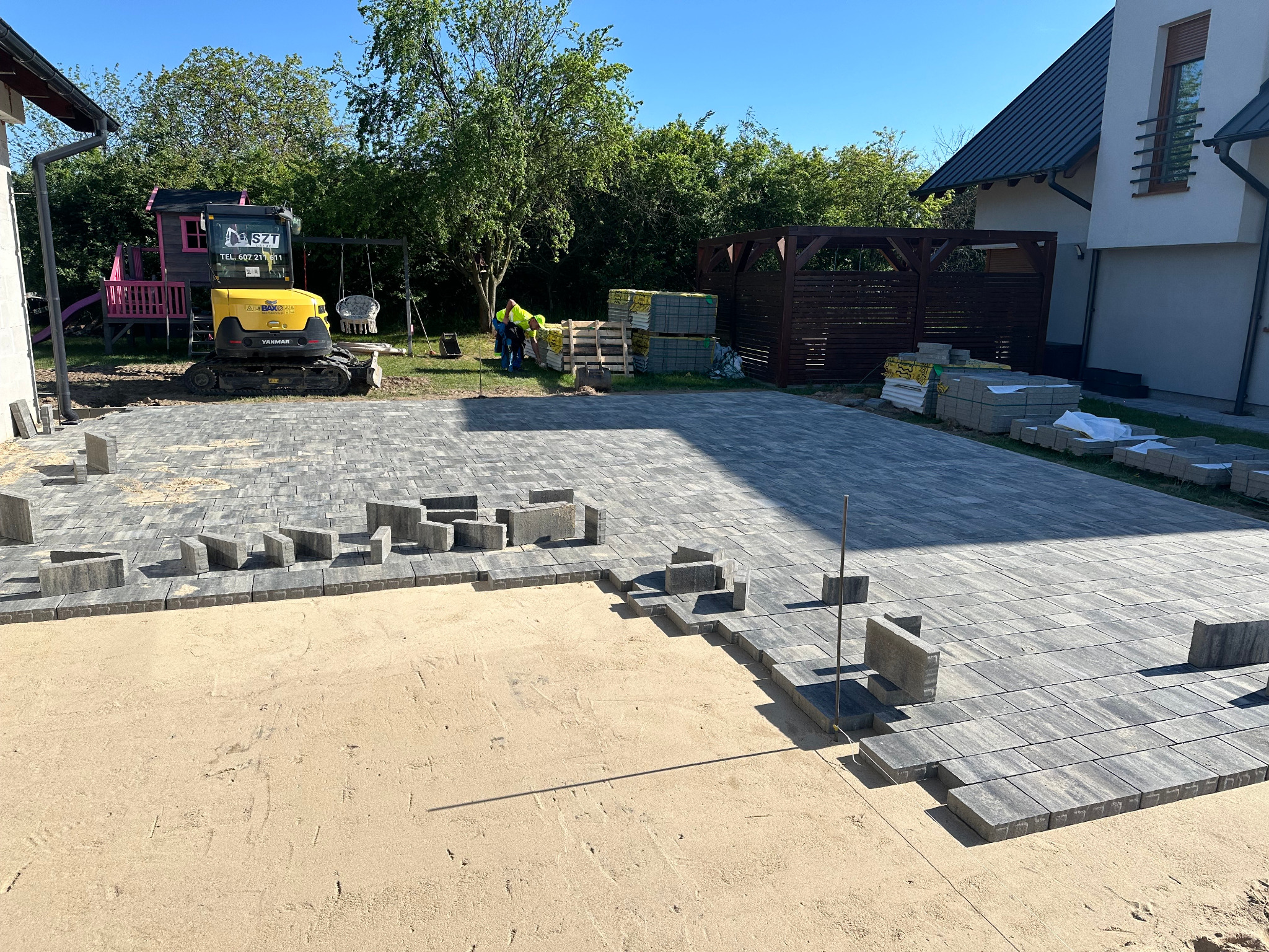 Układanie szarej kostki brukowej na podsypce piaskowej. W tle minikoparka, palety z kostką i budynek mieszkalny. Widok z góry, w słoneczny dzień.