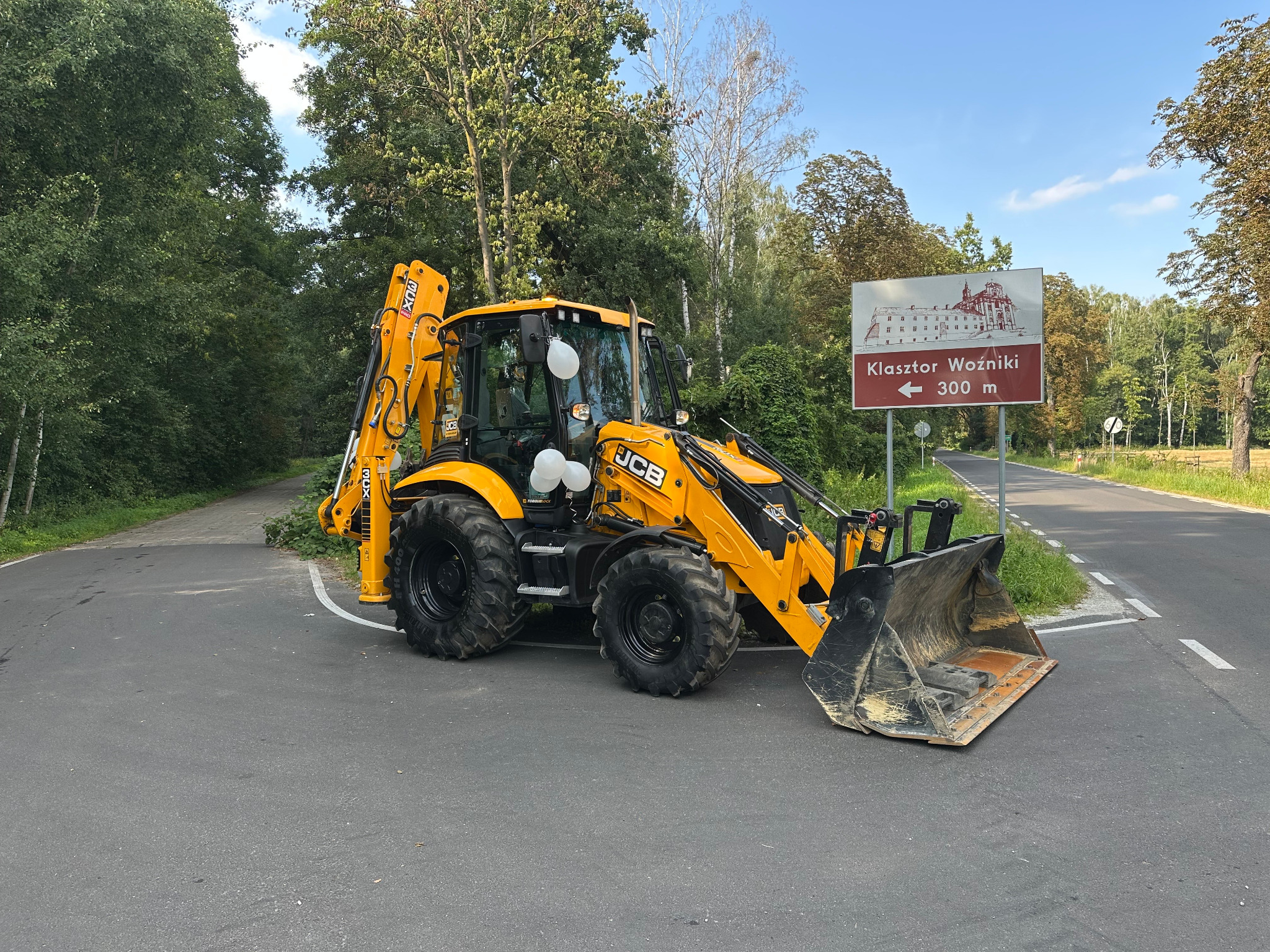 Żółta koparko-ładowarka JCB zaparkowana na asfalcie, obok drogowskazu na Klasztor Woźniki. Maszyna z białymi balonami, otoczona zielenią drzew.