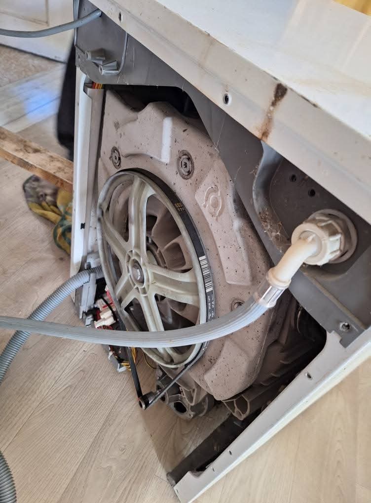 Wnętrze pralki automatycznej z widocznym bębnem, paskiem napędowym i elementami instalacji wodnej. Widoczne ślady użytkowania i zabrudzenia.