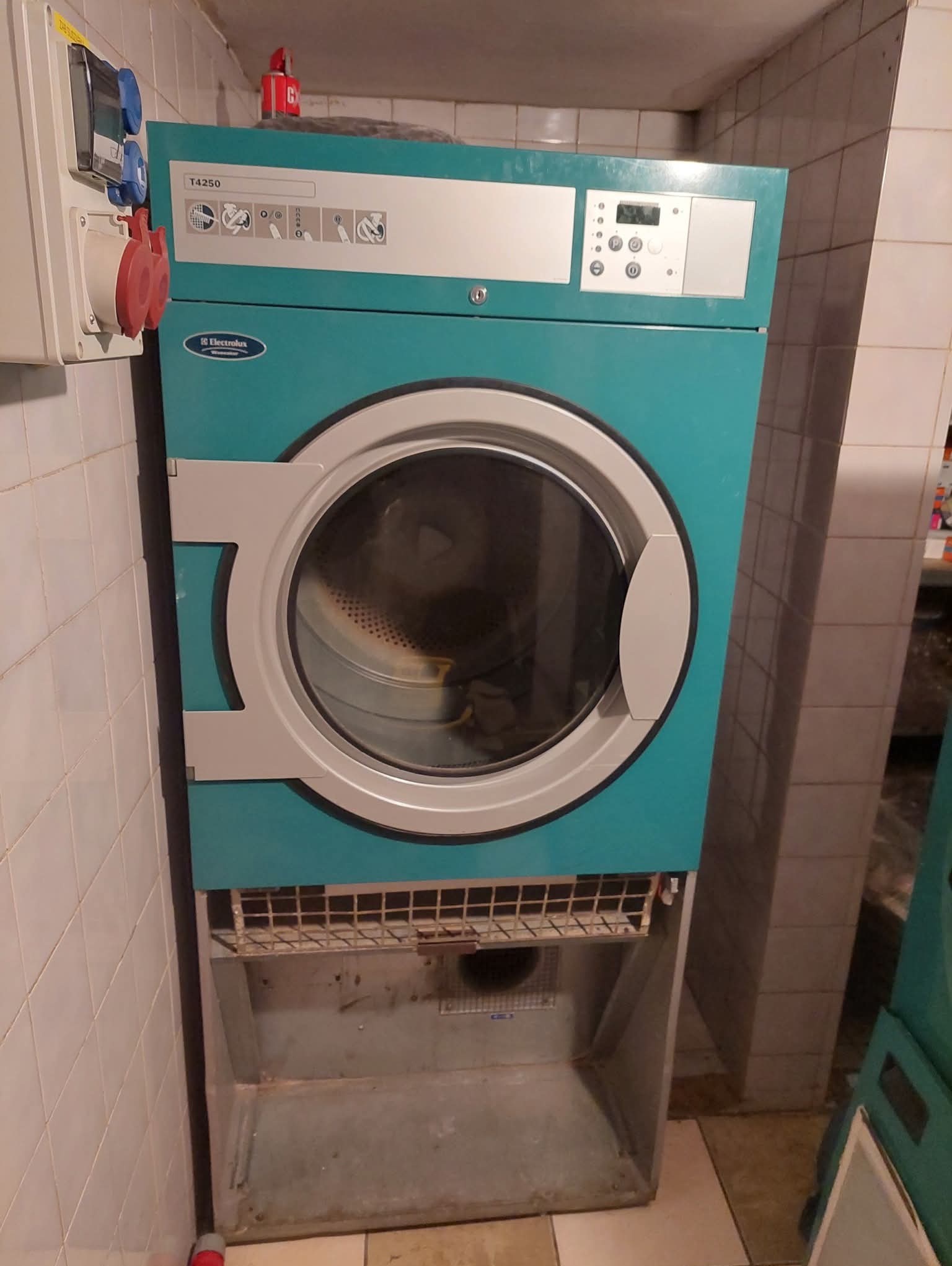 Profesjonalna suszarka przemysłowa Electrolux T4250 w pralni, z widocznymi panelami sterowania i gniazdami zasilania. Tło stanowią kafelki.