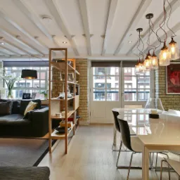 Salon w stylu loft z ceglaną ścianą, drewnianymi belkami na suficie, nowoczesnym oświetleniem w postaci szklanych słoików zawieszonych na kablach i białym stołem z czarnymi krzesłami.