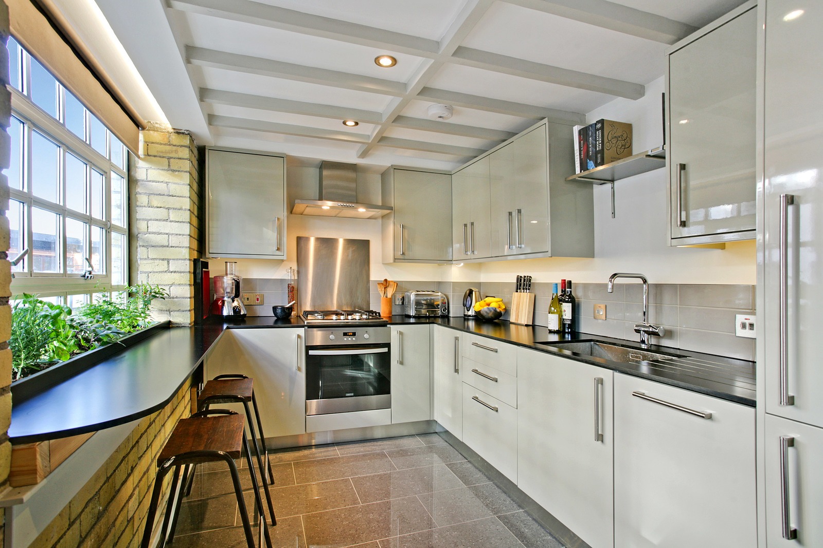 Nowoczesna, jasna kuchnia z białymi szafkami, czarnym blatem, stalowym okapem, oknem z ziołami i drewnianymi stołkami barowymi.