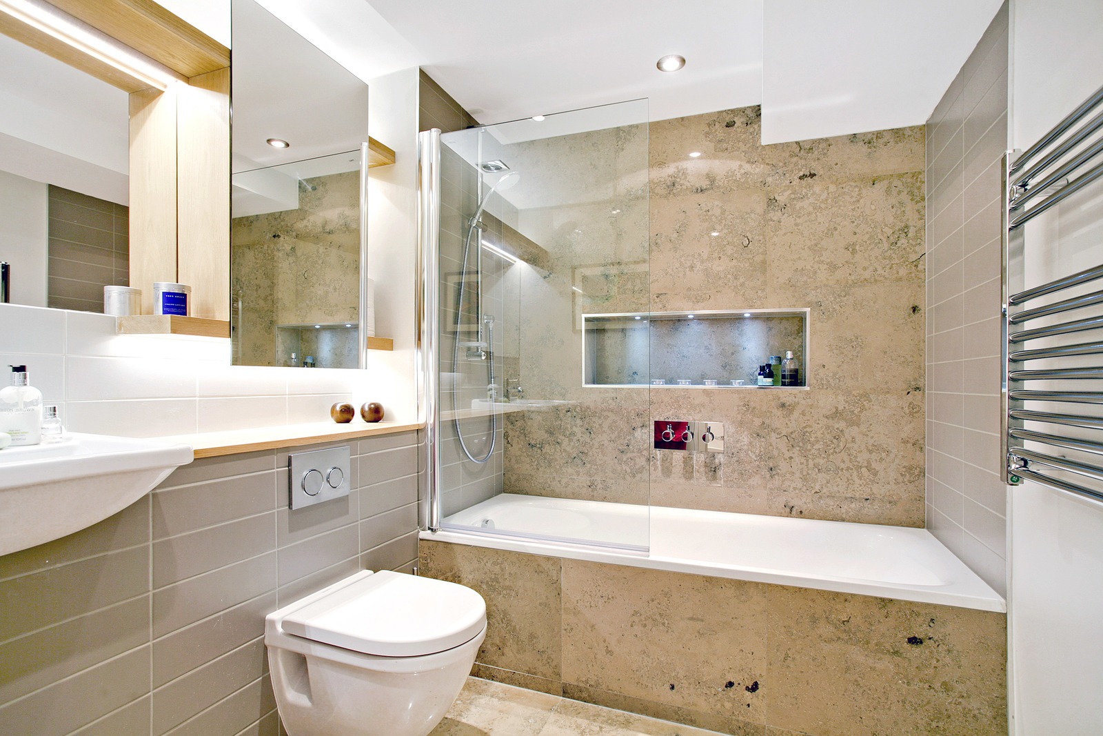 Nowoczesna łazienka z beżowymi płytkami imitującymi kamień, białą wanną z parawanem, umywalką, toaletą i grzejnikiem drabinkowym.
