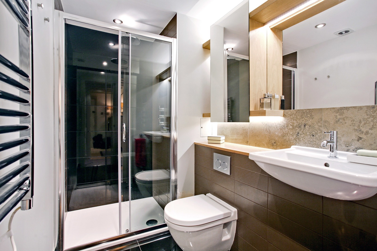 Nowoczesna łazienka z kabiną prysznicową z przesuwnymi drzwiami, podwieszaną toaletą, umywalką i podświetlanym lustrem, wykończona brązowymi płytkami i elementami drewnianymi.