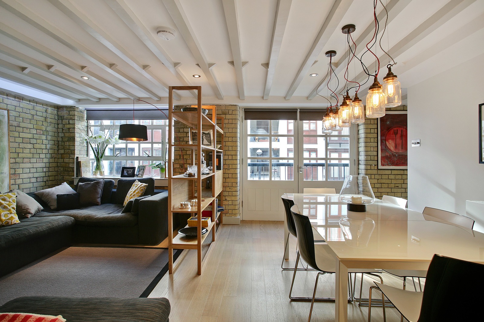 Salon w stylu loft z ceglaną ścianą, drewnianymi belkami na suficie, nowoczesnym oświetleniem w postaci szklanych słoików zawieszonych na kablach i białym stołem z czarnymi krzesłami.