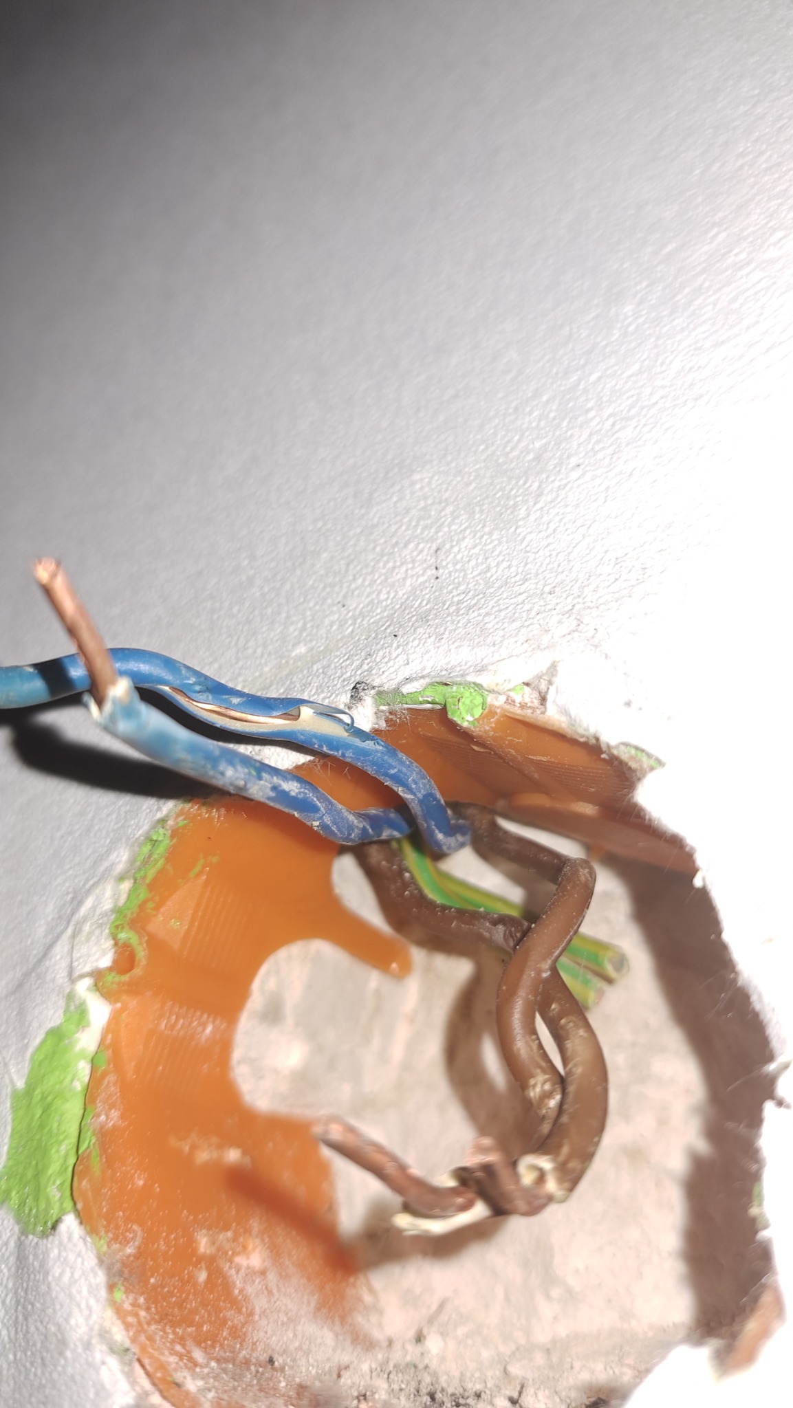 Fragment ściany z wystającymi przewodami elektrycznymi w kolorach niebieskim i brązowym, otoczonymi resztkami gipsu i zielonej farby, widoczne niedociągnięcia w wykończeniu.