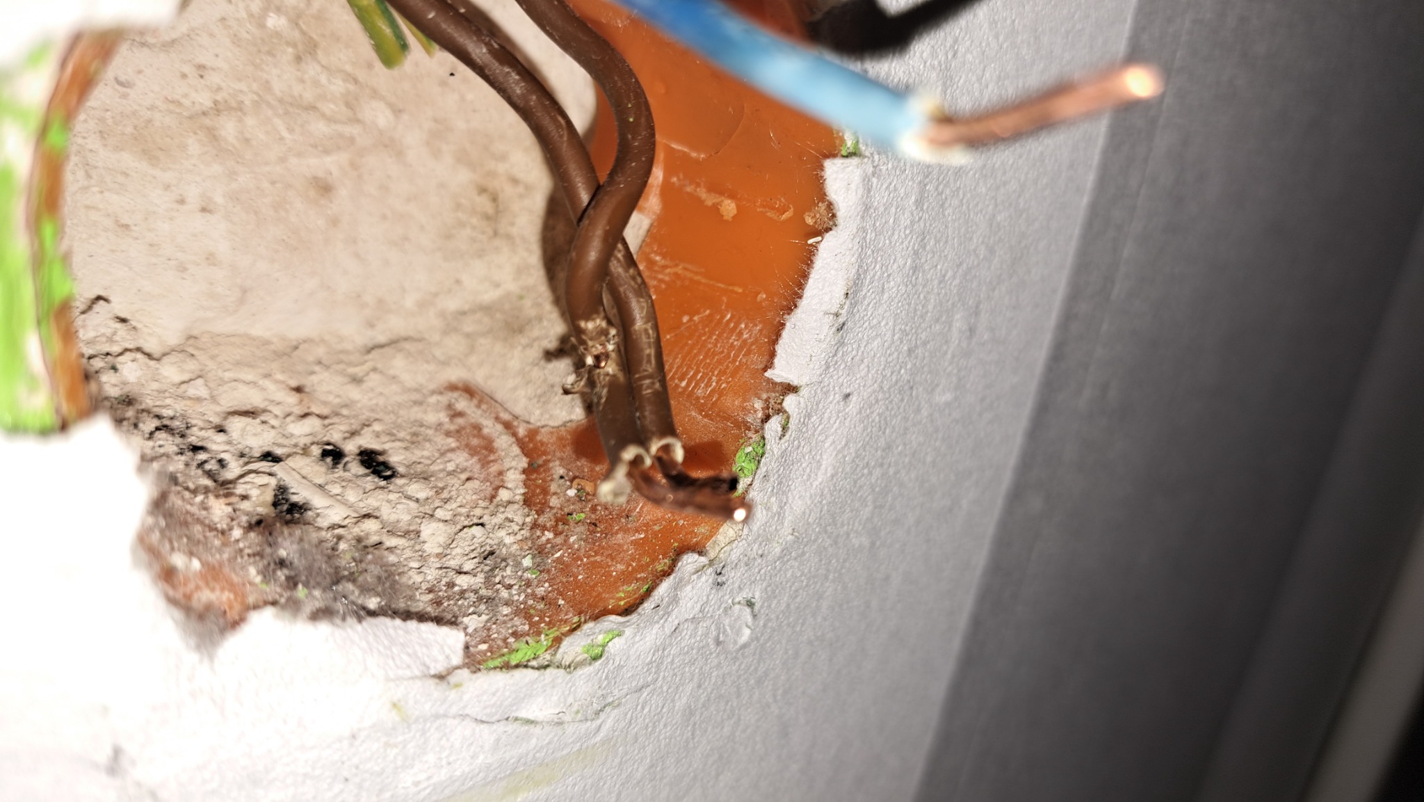 Widok z bliska na niechlujnie przygotowane gniazdo elektryczne z wystającymi przewodami w kolorach brązowym i niebieskim, osadzone w pomarańczowej puszce w ścianie z widocznymi śladami gipsu i farby.