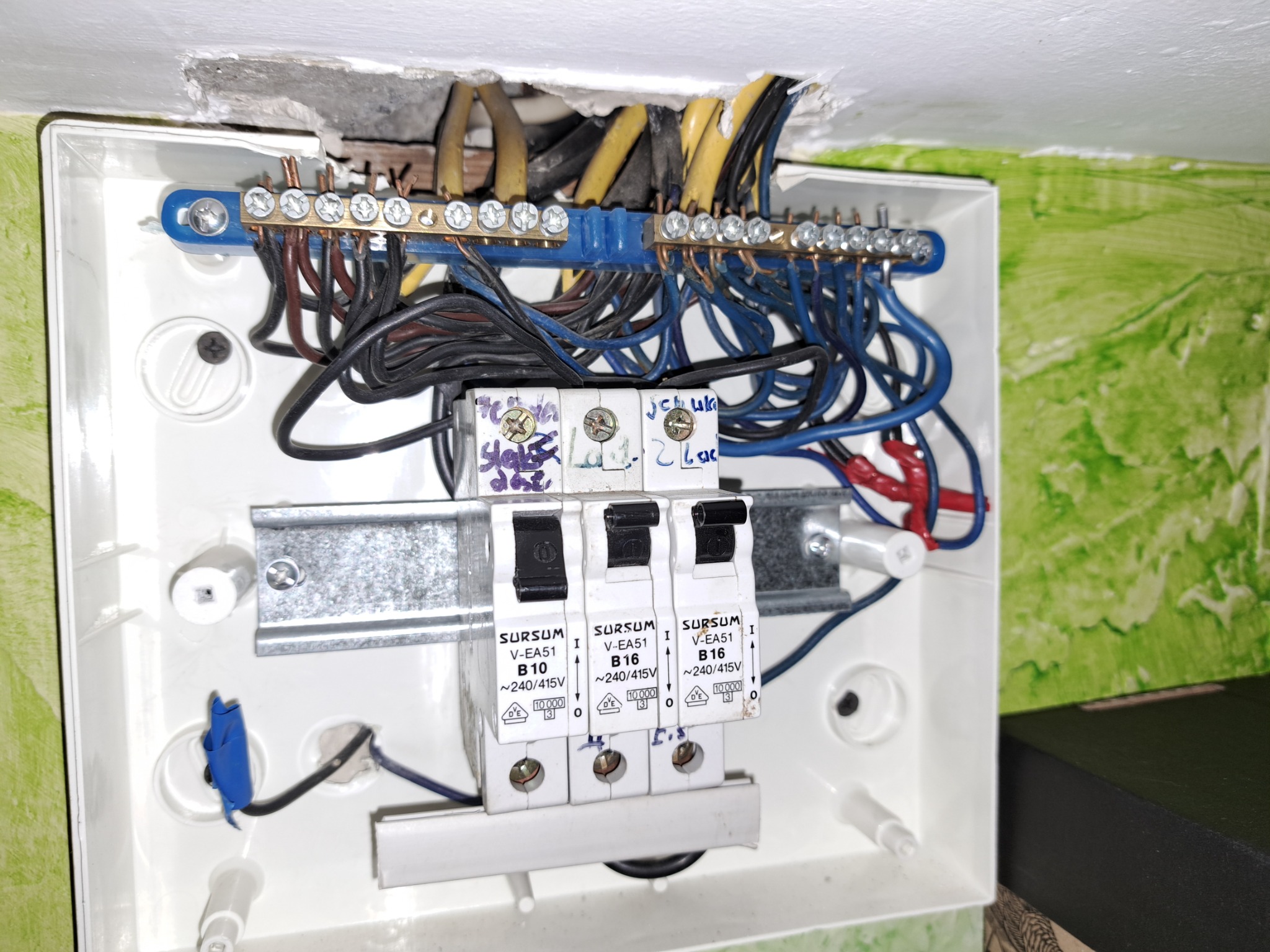 Otwarta skrzynka elektryczna z widocznymi bezpiecznikami Sursum, przewodami w różnych kolorach i listwami zaciskowymi, zamontowana na ścianie z zieloną tapetą i uszkodzonym tynkiem powyżej.