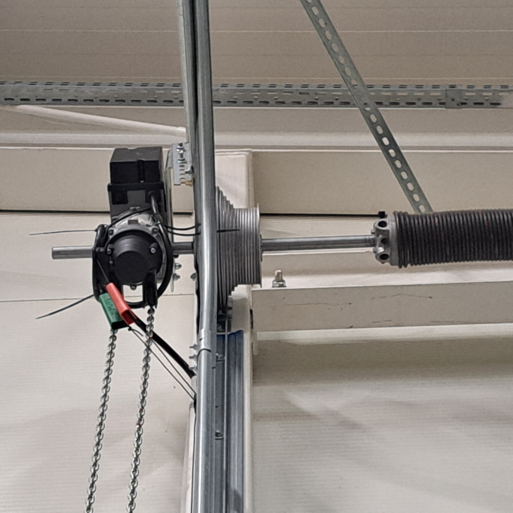 Mechanizm napędu bramy garażowej z silnikiem, łańcuchem i sprężyną skrętną, widoczny na tle białej ściany i sufitu.