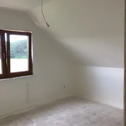 Poddasze po pracach wykończeniowych: białe ściany, okno z widokiem na las, wystający kabel oświetleniowy z sufitu, surowa podłoga przygotowana do dalszych prac.