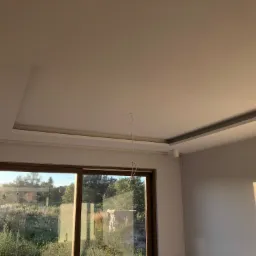 Wykończony sufit z wnęką i punktowym oświetleniem, widok na zewnątrz przez duże okno przesuwne, widoczne przewody oświetleniowe.