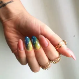 Dłoń z manicure hybrydowym w odcieniach granatu i żółci, zdobionym białymi gwiazdkami i złotą linią, uzupełniona złotymi pierścionkami z cyrkoniami, na tle jasnej powierzchni.