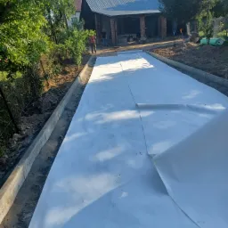 Przygotowanie podłoża pod nawierzchnię: rozłożona biała geowłóknina pomiędzy betonowymi obrzeżami, w tle widoczni pracownicy i budynek gospodarczy.