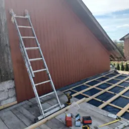 Prace przygotowawcze do ocieplenia poddasza: widoczna drabina oparta o ścianę budynku, rozłożona folia izolacyjna i drewniane łaty, narzędzia pracy, w tym wkrętarki, młotek i poziomice.