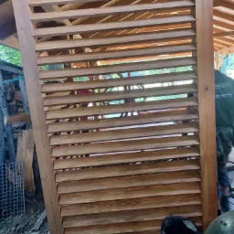 Drewniana konstrukcja altany z lamelami, widoczny fragment więźby dachowej, element roboczy w tle, torba narzędziowa na betonowej podstawie.