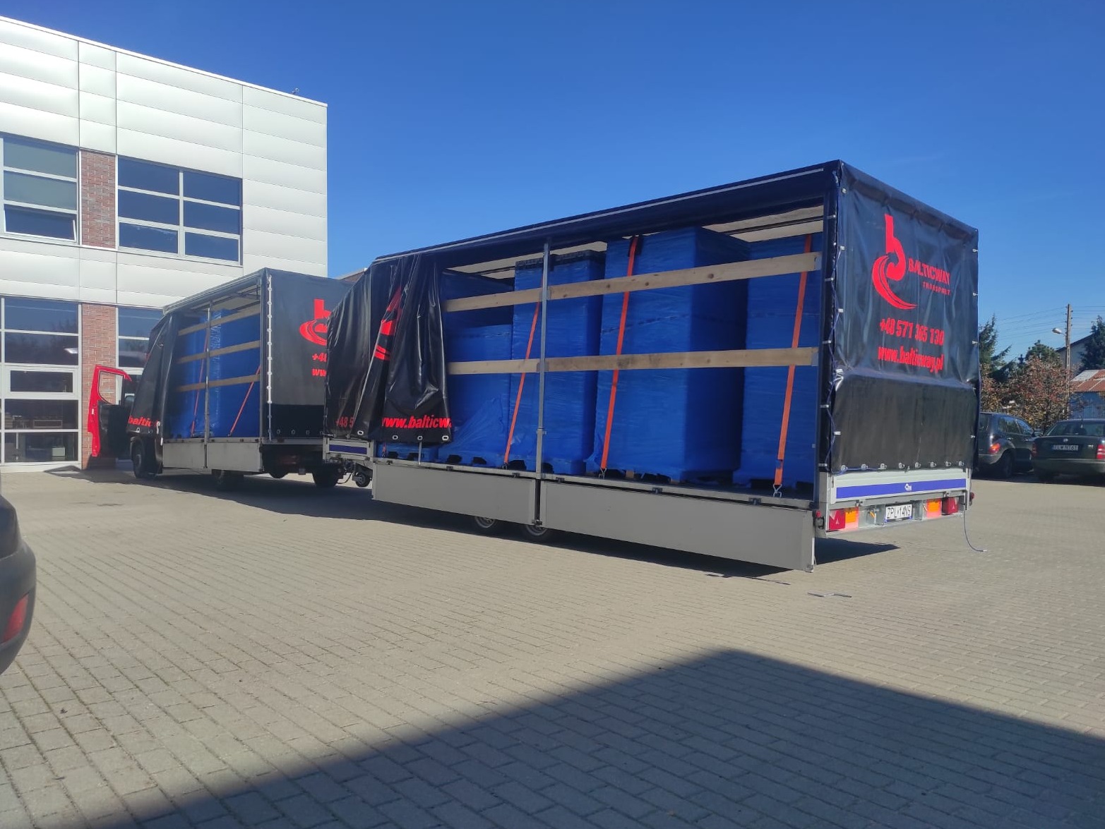 Dwa busy z naczepami, z czarnymi plandekami z logo firmy, załadowane niebieskimi pakunkami, zabezpieczonymi pomarańczowymi pasami, stoją na brukowanym placu przed budynkiem w słoneczny dzień.