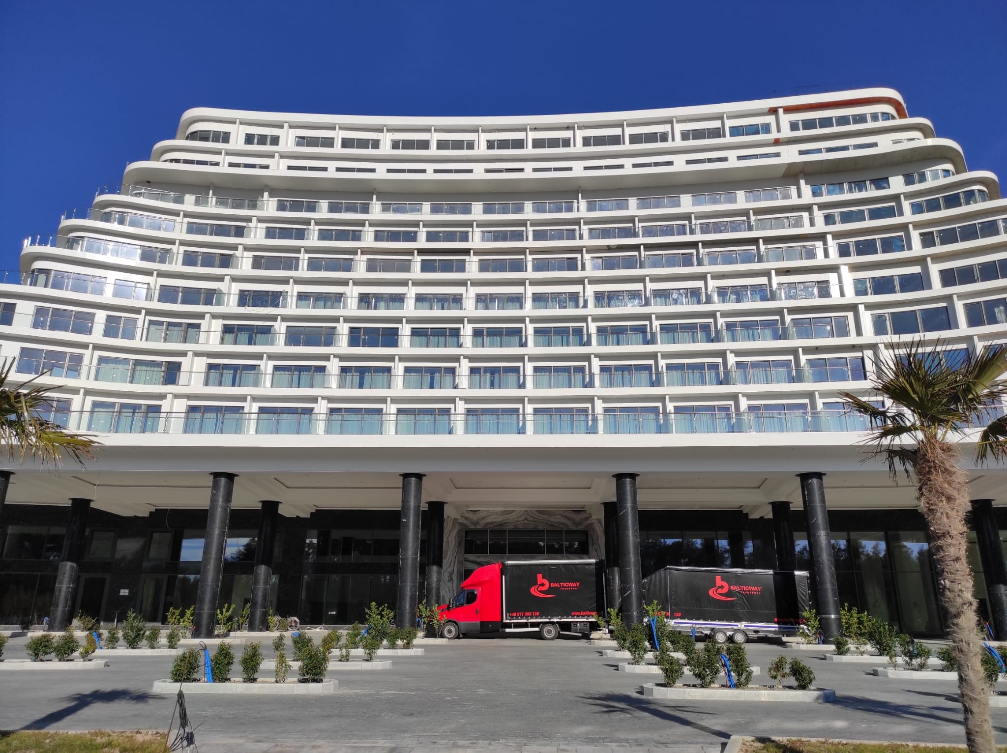 Dwa busy z logo firmy transportowej Balticway zaparkowane przed nowoczesnym, białym budynkiem hotelowym z licznymi balkonami i palmami, w słoneczny dzień.