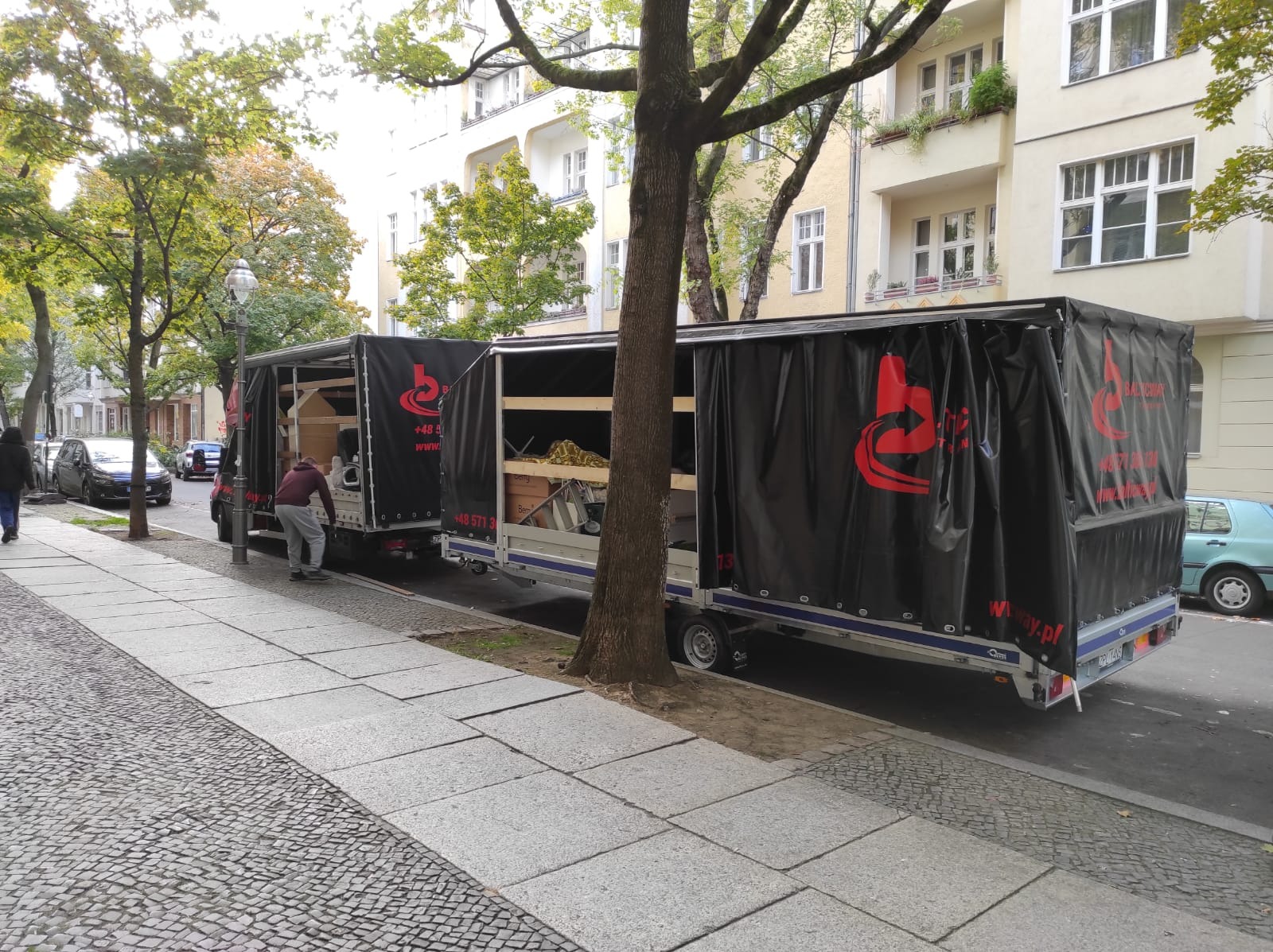 Dwa przyczepiane kontenery transportowe z czarną plandeką i czerwonym logo firmy, zaparkowane na ulicy w mieście, mężczyzna w szarym dresie ładuje kartony do pierwszego kontenera.