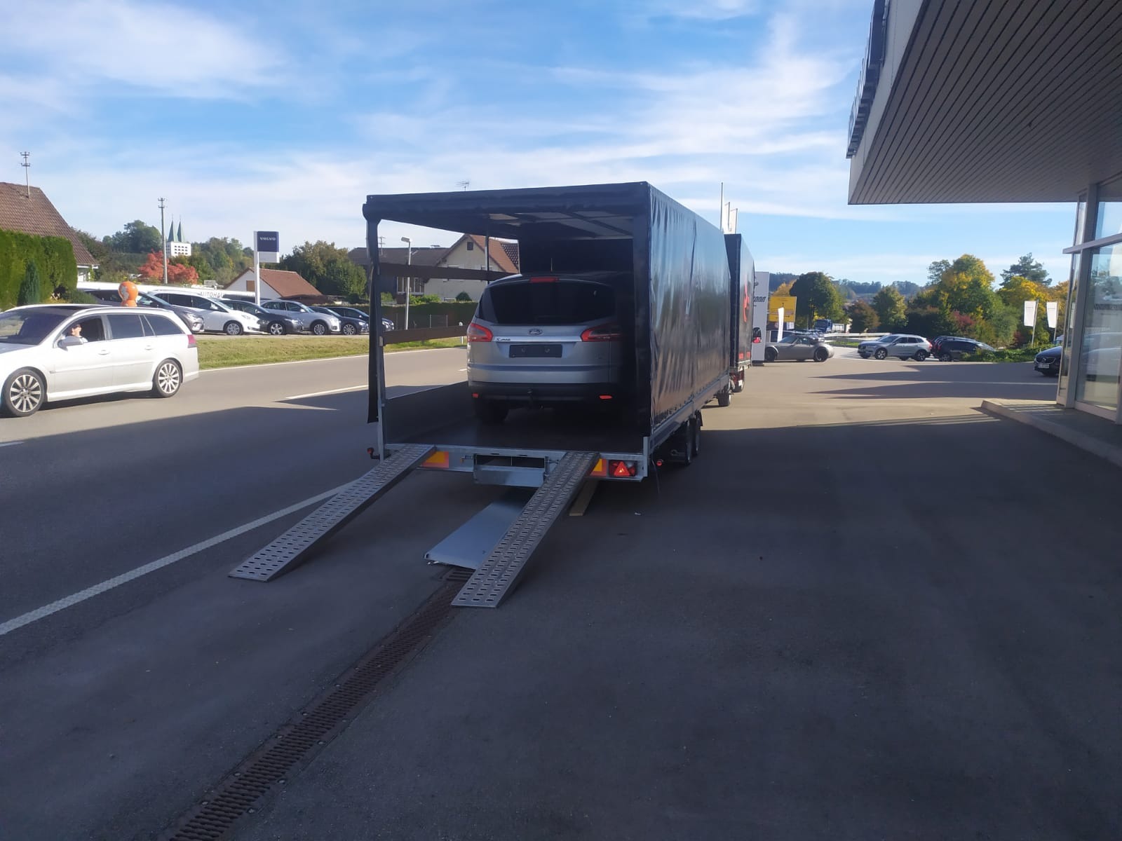 Srebrny samochód osobowy na platformie lawety z rozłożonymi najazdami, widok z tyłu, w tle parking z innymi pojazdami i budynki.