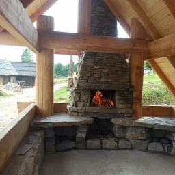 Kamienny kominek zewnętrzny z palącym się ogniem, drewniana konstrukcja altany w tle, widok na wiejski krajobraz.