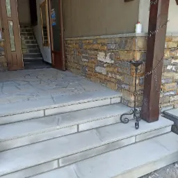 Kamienne schody wejściowe z jasnoszarego kamienia prowadzące do domu, z elementami dekoracyjnymi: metalowym świecznikiem i małym drzewkiem iglastym w czarnej doniczce. Ściana obłożona kamieniem...