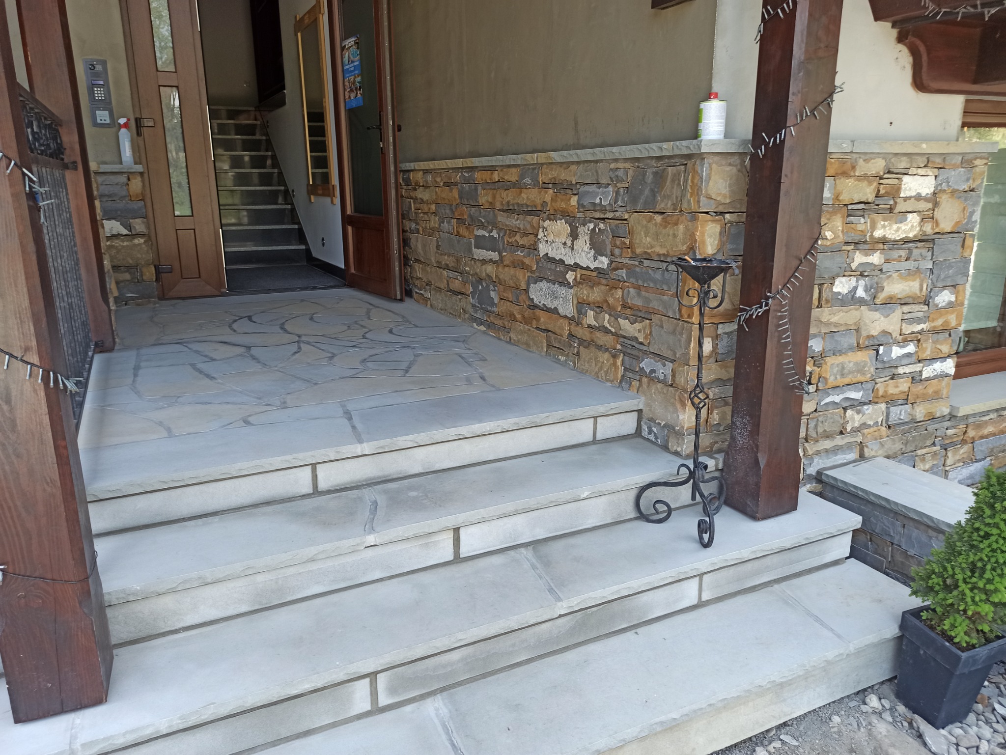 Kamienne schody wejściowe z jasnoszarego kamienia prowadzące do domu, z elementami dekoracyjnymi: metalowym świecznikiem i małym drzewkiem iglastym w czarnej doniczce. Ściana obłożona kamieniem...