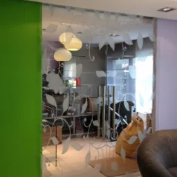 Szklane drzwi przesuwne z matowym wzorem roślinnym, widoczne oświetlenie sufitowe, zielona ściana po lewej stronie i fragment szarego fotela po prawej.