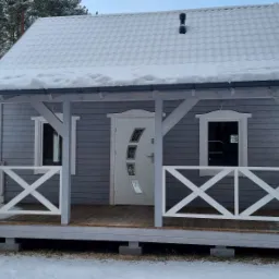 Mały, szary domek z drewnianą werandą i śniegiem na dachu, otoczony zimowym lasem.