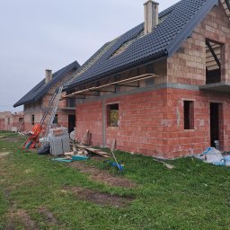 USŁUGI OGÓLNOBUDOWLANE PAWEŁ HARAF - Budowa domu z czerwonej cegły: wzniesione ściany, częściowo położony dach z czarnej dachówki. Na pierwszym planie trawa, w tle kolejne budynki w trakcie budowy.
