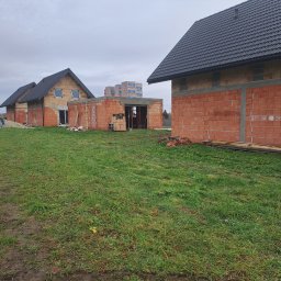 USŁUGI OGÓLNOBUDOWLANE PAWEŁ HARAF - Budowa domów jednorodzinnych w stanie surowym otwartym z czerwonej cegły. Widoczne ściany, dachy z czarnej dachówki i palety z materiałami budowlanymi.