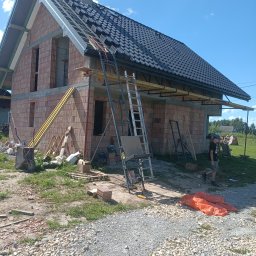 USŁUGI OGÓLNOBUDOWLANE PAWEŁ HARAF - Budowa domu z czerwonej cegły, widoczne rusztowania, drabiny i częściowo ułożony dach z czarnej dachówki. Na pierwszym planie gruz i chłopak w tle.