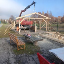 USŁUGI OGÓLNOBUDOWLANE PAWEŁ HARAF - Montaż drewnianej konstrukcji z użyciem dźwigu samochodowego. Widoczny pracownik w pomarańczowym ubraniu. W tle drzewa i niebieskie niebo.