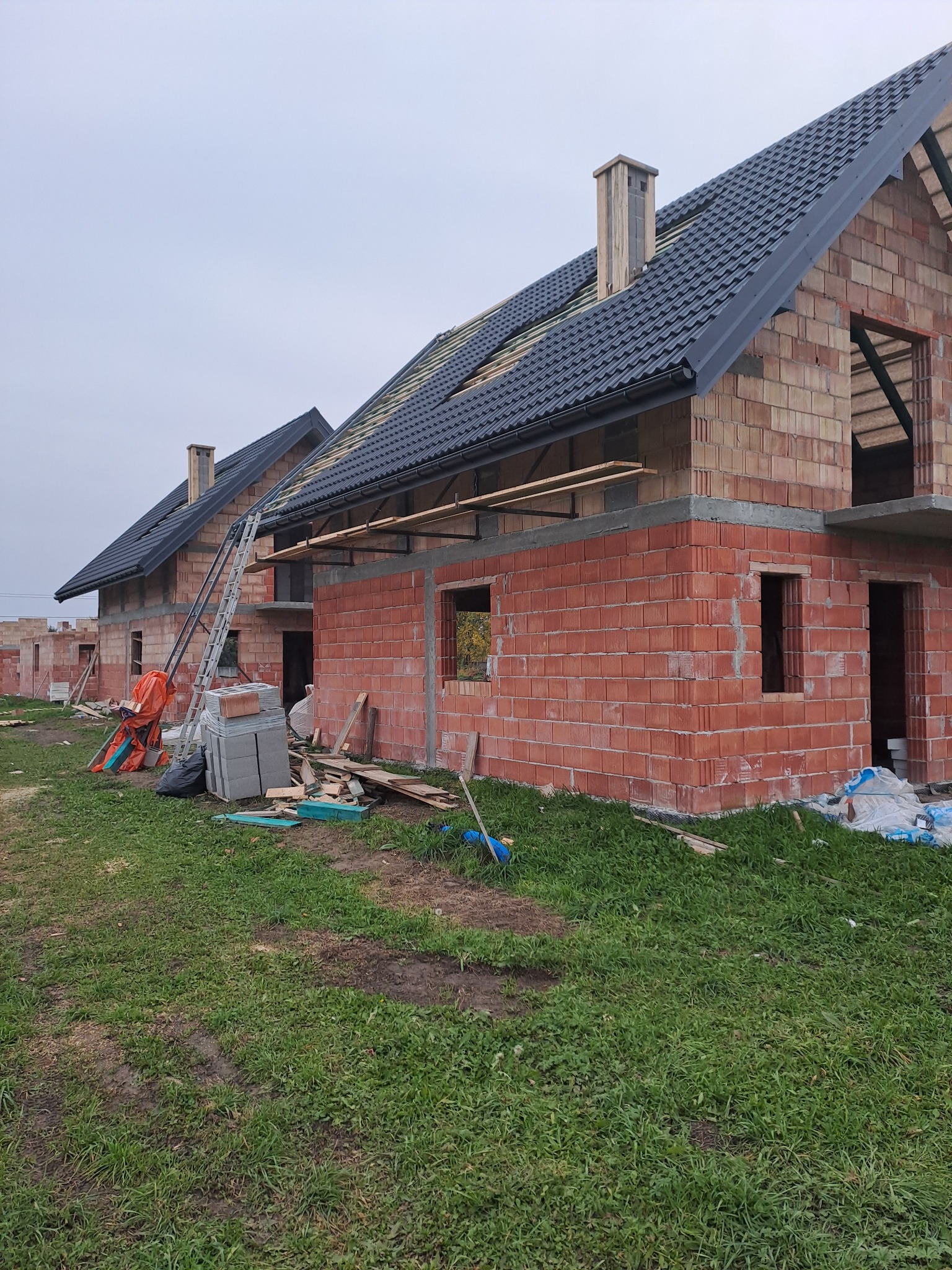 Budowa domu z czerwonej cegły: wzniesione ściany, częściowo położony dach z czarnej dachówki. Na pierwszym planie trawa, w tle kolejne budynki w trakcie budowy.